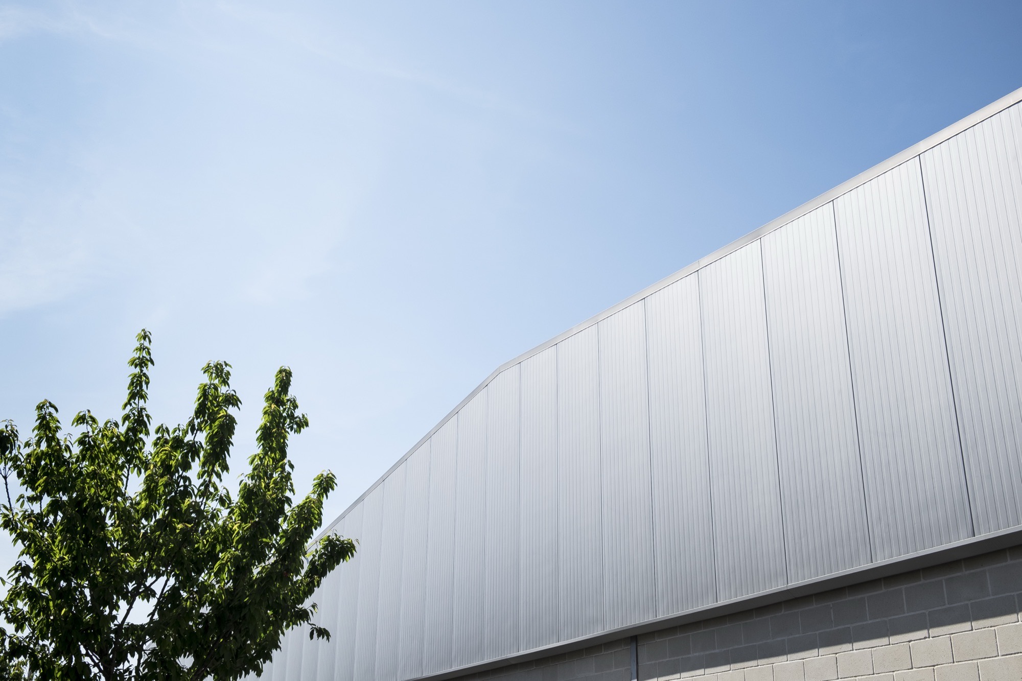 Exterior of the JVperfis building: a silver profiled-sheet wall rising on the diagonal against a clear blue sky, a green tree at the lower left and a concrete block base.