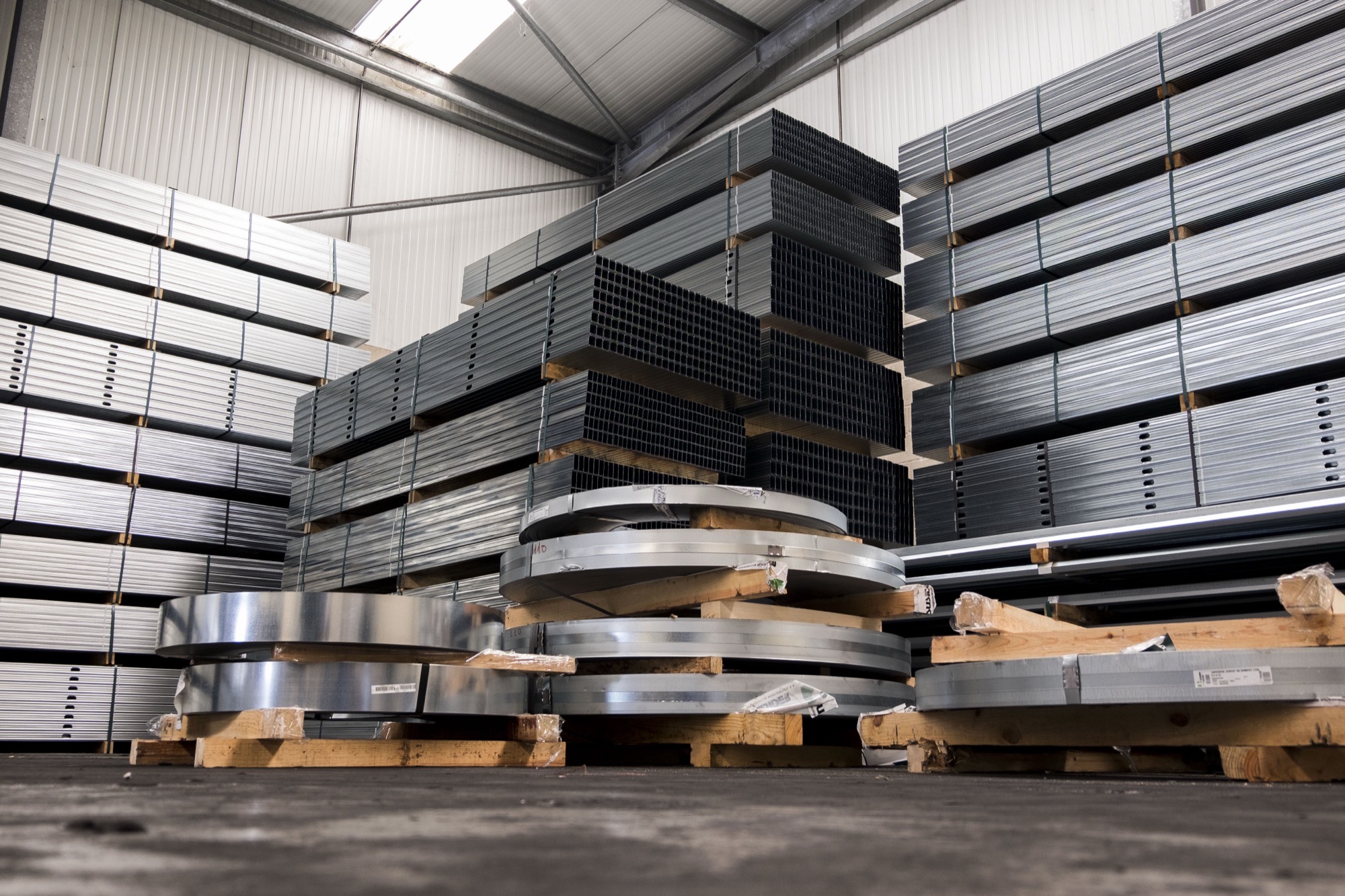 Low-angle view in the warehouse: towers of steel profiles stacked on timber pallets, sheet coils in the foreground and a metal roof above.