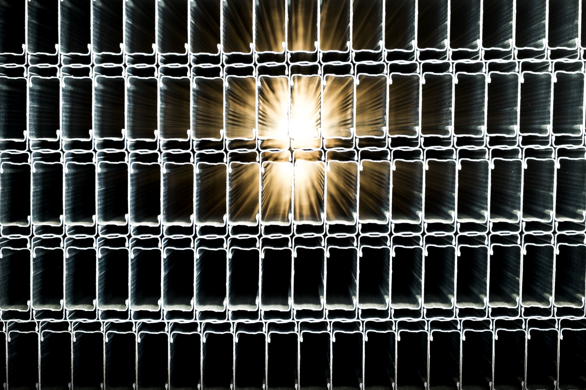 Backlit grid of stud cross-sections stacked together, with the sun bursting in a star through two openings in the dark metal grid.