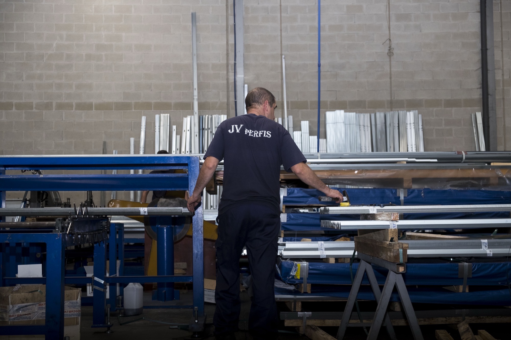 A worker seen from behind, in a JV PERFIS shirt, at a blue production bench surrounded by cut steel profiles, beside a blockwork wall.
