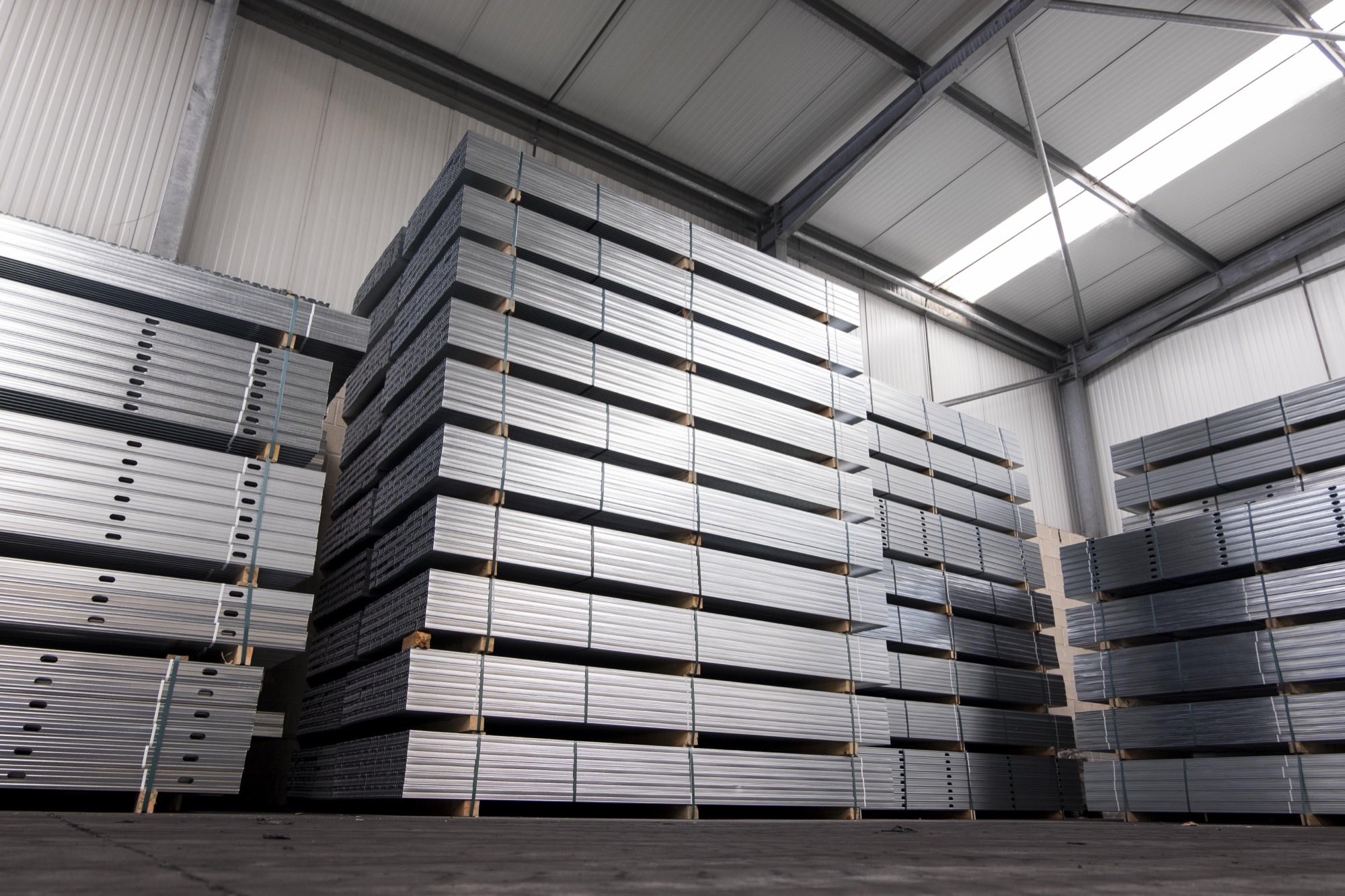Warehouse lit by skylights, with large ordered stacks of steel profiles resting on timber pallets.